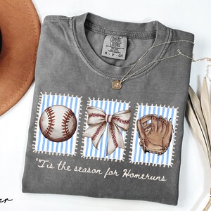 May include: Gray Comfort Colors t-shirt with baseball-themed graphics. The shirt features a baseball, a bow, and a baseball glove on blue and white striped backgrounds. The text "'Tis the season for Homeruns" is printed below the graphics. A brown hat and necklace are also visible.