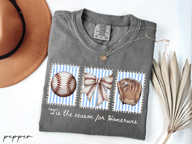 May include: Gray Comfort Colors t-shirt with baseball-themed graphics. The shirt features a baseball, a bow, and a baseball glove on blue and white striped backgrounds. The text "'Tis the season for Homeruns" is printed below the graphics. A brown hat and necklace are also visible.