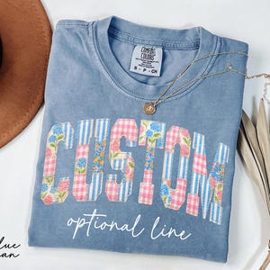 May include: A blue jean colored t-shirt with the word "CUSTOM" in a collegiate style font. The letters are decorated with a mix of floral, gingham, and striped patterns. The text "optional line" is below the word "CUSTOM". A gold necklace is draped across the shirt.