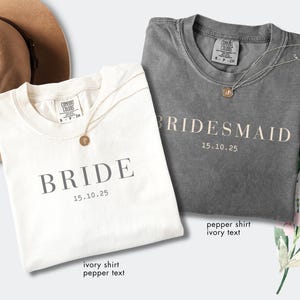 May include: Two t-shirts, one ivory and one pepper, are displayed. The ivory shirt reads "BRIDE" with the date "15.10.25" in a gray font. The pepper shirt says "BRIDESMAID" with the same date. Both shirts have a gold necklace.