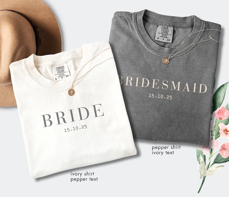 May include: Two t-shirts, one ivory and one pepper, are displayed. The ivory shirt reads "BRIDE" with the date "15.10.25" in a gray font. The pepper shirt says "BRIDESMAID" with the same date. Both shirts have a gold necklace.
