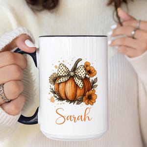 May include: White ceramic mug with a black handle and rim, featuring an orange pumpkin design with a polka dot bow and floral accents. The name "Sarah" is written in an orange script font. A fall-themed mug.