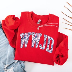 May include: A red sweatshirt with the letters "WWJD" in a floral design. The sweatshirt is paired with blue jeans and a silver necklace. The word "red" is written in the bottom right corner.
