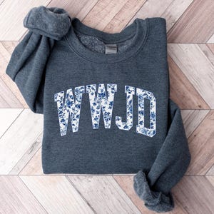 May include: A dark gray sweatshirt with the letters "WWJD" in a white, floral design. The floral pattern features blue flowers and leaves. The sweatshirt is folded on a light wood-grain surface.