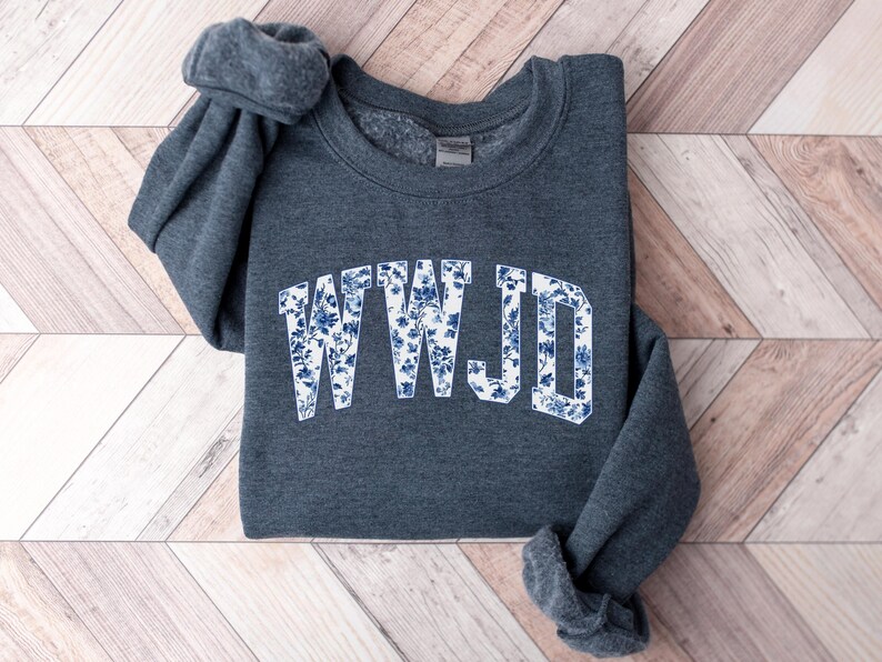 May include: A dark gray sweatshirt with the letters "WWJD" in a white, floral design. The floral pattern features blue flowers and leaves. The sweatshirt is folded on a light wood-grain surface.