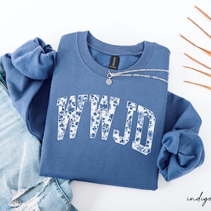 folded indigo blue sweatshirt with large floral WWJD letters, styled with denim and jewelry, soft Christian gift for women
