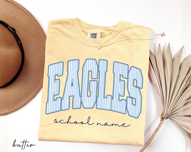 K&ouml;nnte beinhalten: Ein buttergelbes Kurzarm-T-Shirt mit dem Wort EAGLES in gro&szlig;en, blau-wei&szlig; vertikal gestreiften Blockbuchstaben mit blauem Umriss. Unter dem Wort EAGLES ist der Text school name in einer dunkelblauen Schreibschrift aufgedruckt.