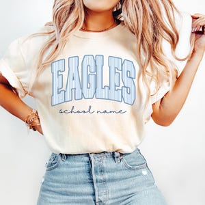 K&ouml;nnte beinhalten: Ein cremefarbenes Kurzarm-T-Shirt mit dem Wort EAGLES in gro&szlig;en, blau-wei&szlig; gestreiften Buchstaben im College-Stil auf der Brust, wobei die W&ouml;rter school name darunter in einer kleineren, dunkelblauen Schreibschrift zu sehen sind.