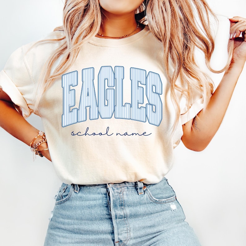 K&ouml;nnte beinhalten: Ein cremefarbenes Kurzarm-T-Shirt mit dem Wort EAGLES in gro&szlig;en, blau-wei&szlig; gestreiften Buchstaben im College-Stil auf der Brust, wobei die W&ouml;rter school name darunter in einer kleineren, dunkelblauen Schreibschrift zu sehen sind.
