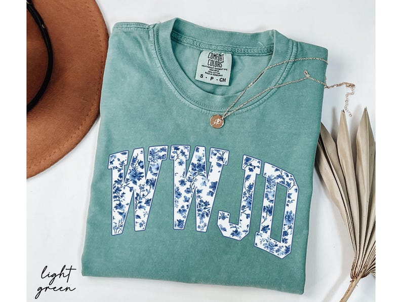 May include: Light green t-shirt with the letters "WWJD" in a blue floral pattern. A gold necklace with a pendant and a brown hat are also visible. The shirt is folded and has a Comfort Colors tag.