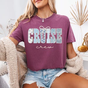 Puede incluir: Una camiseta granate con la palabra "CRUISE" en letras grandes con dise&ntilde;os en azul claro y blanco. La palabra "crew" est&aacute; escrita en cursiva debajo. La camiseta es de manga corta y tiene cuello redondo.