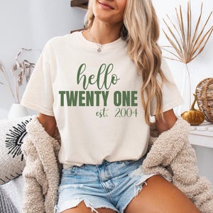 Op de afbeelding: Cr&egrave;mekleurig T-shirt met de tekst "hello TWENTY ONE est. 2004" in het groen. Het shirt heeft een casual stijl, geschikt voor dagelijks gebruik. De afbeelding toont een persoon die het shirt draagt.