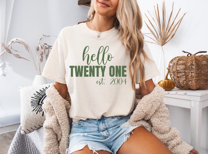 Op de afbeelding: Cr&egrave;mekleurig T-shirt met de tekst "hello TWENTY ONE est. 2004" in het groen. Het shirt heeft een casual stijl, geschikt voor dagelijks gebruik. De afbeelding toont een persoon die het shirt draagt.
