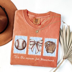 May include: Orange Comfort Colors t-shirt with baseball-themed graphics. The design includes a baseball, a bow, and a baseball glove on blue and white striped backgrounds. The text "'Tis the season for Homeruns" is printed below the graphics.