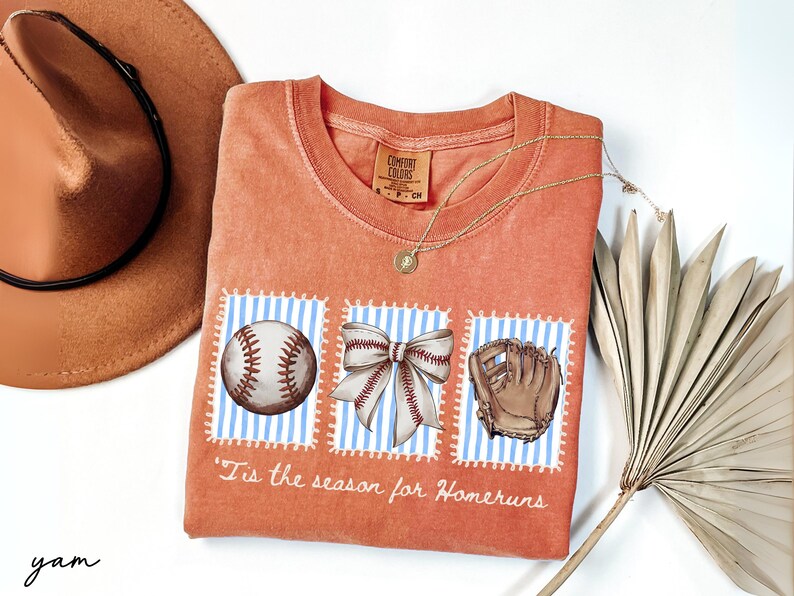 May include: Orange Comfort Colors t-shirt with baseball-themed graphics. The design includes a baseball, a bow, and a baseball glove on blue and white striped backgrounds. The text "'Tis the season for Homeruns" is printed below the graphics.