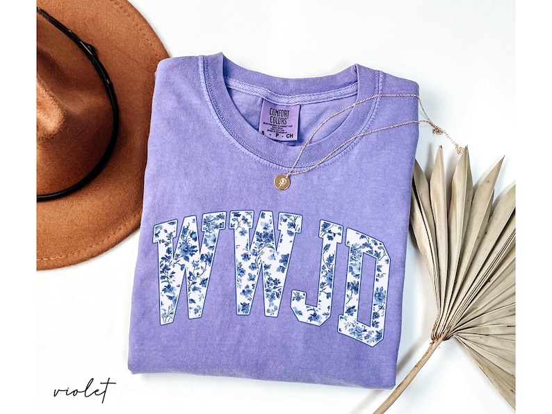May include: A violet Comfort Colors t-shirt with the letters WWJD in a floral design. A gold necklace with a pendant and a brown hat are also in the image. The shirt is a crew neck.