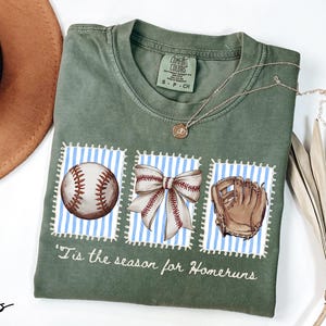 May include: Moss green t-shirt with baseball-themed graphics. The shirt features a baseball, a bow, and a baseball glove on blue and white striped backgrounds. Text on the shirt reads "'Tis the season for Homeruns". A brown hat and necklace are also visible.
