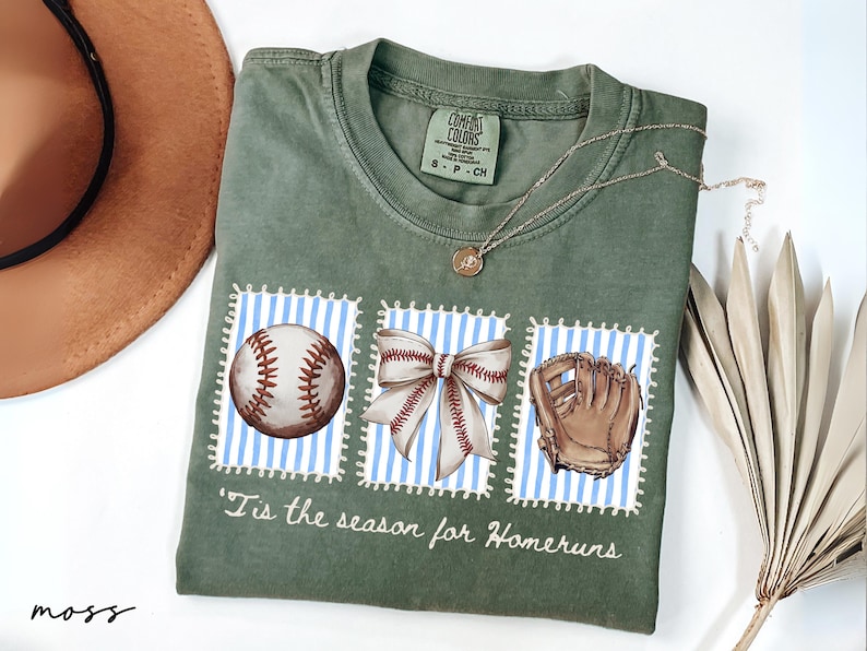 May include: Moss green t-shirt with baseball-themed graphics. The shirt features a baseball, a bow, and a baseball glove on blue and white striped backgrounds. Text on the shirt reads "'Tis the season for Homeruns". A brown hat and necklace are also visible.