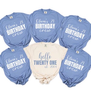 Op de afbeelding: Een verzameling lichtblauwe en cr&egrave;mekleurige t-shirts. De lichtblauwe shirts hebben de tekst "Olivia's 21 BIRTHDAY crew" in het wit. Het cr&egrave;mekleurige shirt zegt "hello TWENTY ONE est. 2004".