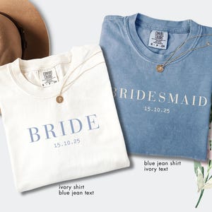 May include: Two t-shirts, one ivory and one blue jean, with the words "BRIDE" and "BRIDESMAID" printed in blue and ivory, respectively. Each shirt has a date printed below the word. Gold necklaces are draped on the shirts. A tan hat is in the background.
