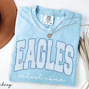 K&ouml;nnte beinhalten: Ein hellblaues Kurzarm-T-Shirt mit Vintage-Waschung. Auf der Vorderseite steht das Wort EAGLES in gro&szlig;en, geschwungenen Blockbuchstaben mit wei&szlig;-blauen Streifen, darunter steht school name in einer wei&szlig;en Schreibschrift.
