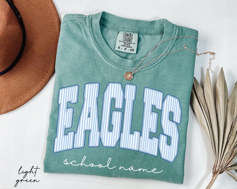 K&ouml;nnte beinhalten: Ein hellgr&uuml;nes Kurzarm-T-Shirt mit Rundhalsausschnitt. Auf der Vorderseite steht das Wort EAGLES in gro&szlig;en, wei&szlig;-blau gestreiften Blockbuchstaben mit blauem Umriss, darunter der Text school name in wei&szlig;er Schreibschrift.