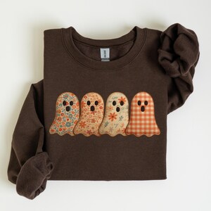 May include: A brown sweatshirt featuring four ghost appliqués with floral and plaid patterns. The ghosts have round eyes and open mouths. The sweatshirt has long sleeves and a crew neck.