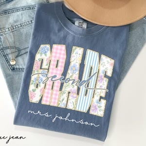 May include: A blue short-sleeve t-shirt featuring the text "GRADE" in large block letters filled with floral, pink gingham, and blue striped patterns. The word "second" is written in cursive over the letters, and "mrs. johnson" is printed below.
