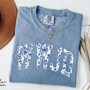 May include: A blue jean Comfort Colors t-shirt with the letters WWJD in a white font filled with a blue floral pattern. A gold necklace with a pendant and a brown hat are also in the image.