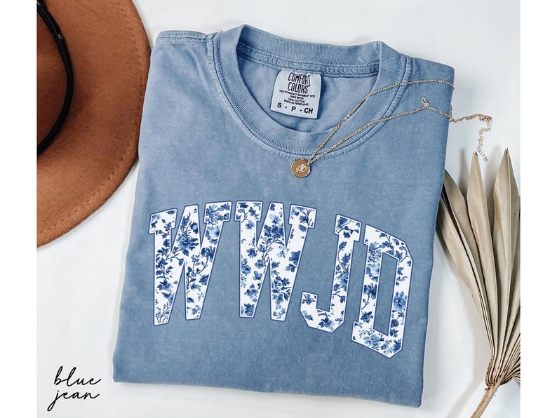 May include: A blue jean Comfort Colors t-shirt with the letters WWJD in a white font filled with a blue floral pattern. A gold necklace with a pendant and a brown hat are also in the image.