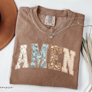 May include: A brown t-shirt with the word "AMEN" appliqued across the front in a floral and checkered pattern. A silver necklace with a small pendant rests on the shirt. A brown hat is visible in the upper left corner.