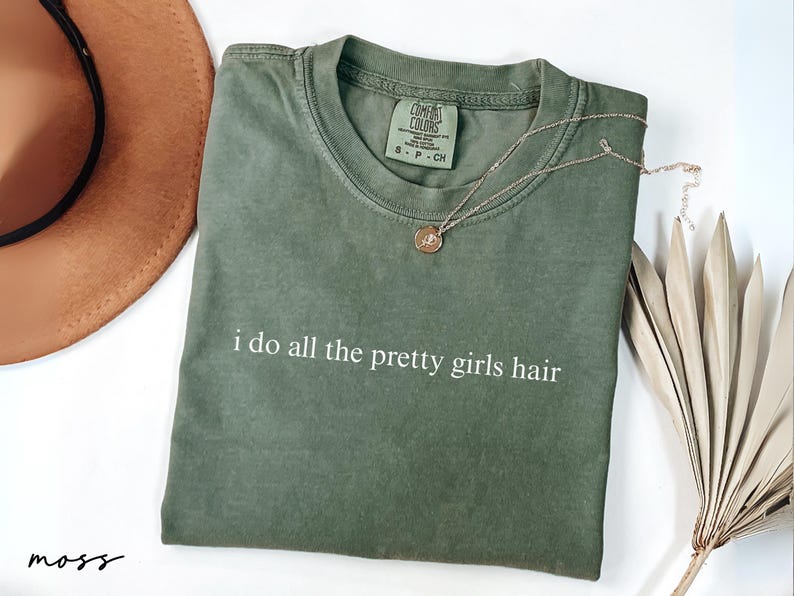 Może przedstawiać: Zielona koszulka w kolorze mchu z białym napisem "i do all the pretty girls hair". Złoty naszyjnik z wisiorkiem spoczywa na koszulce. Widoczny jest r&oacute;wnież brązowy kapelusz i suszone liście palmowe.