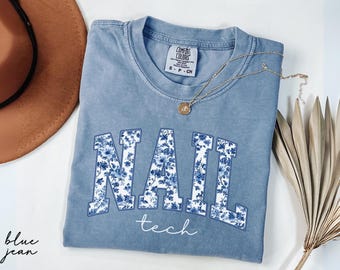 May include: A blue jean colored t-shirt with the words "NAIL tech" in a floral design. The shirt has a crew neck and short sleeves. A gold necklace with a pendant is draped across the shirt. A brown hat and dried plant are in the background.