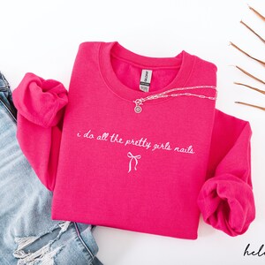 May include: A hot pink sweatshirt with the text "i do all the pretty girls nails" in white script. A silver necklace with a pendant is draped over the shirt. Light blue jeans are visible on the left.