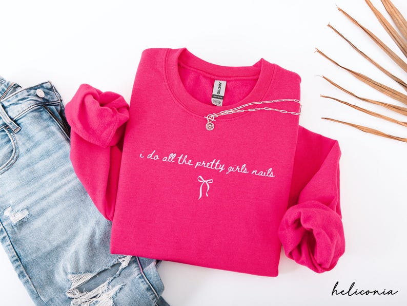May include: A hot pink sweatshirt with the text "i do all the pretty girls nails" in white script. A silver necklace with a pendant is draped over the shirt. Light blue jeans are visible on the left.