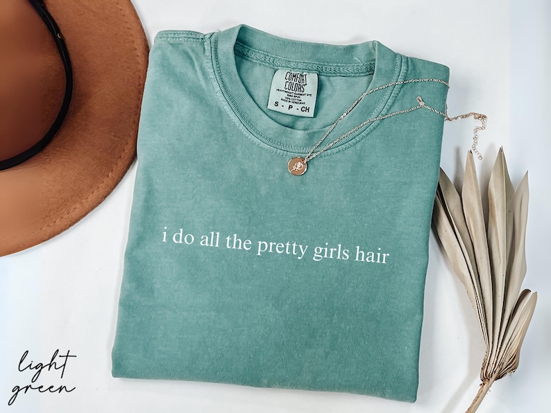 Może przedstawiać: Jasnozielona koszulka Comfort Colors z białym napisem &bdquo;i do all the pretty girls hair&rdquo;. Złoty naszyjnik z wisiorkiem spoczywa na koszulce. Na zdjęciu znajduje się r&oacute;wnież brązowy kapelusz i suszona roślina.