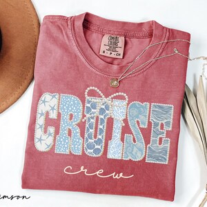 Puede incluir: Camiseta de color carmes&iacute; con las palabras "CRUISE crew". La palabra "CRUISE" es de color azul claro con un patr&oacute;n de conchas marinas y estrellas de mar. Un collar dorado con un colgante est&aacute; sobre la camiseta.