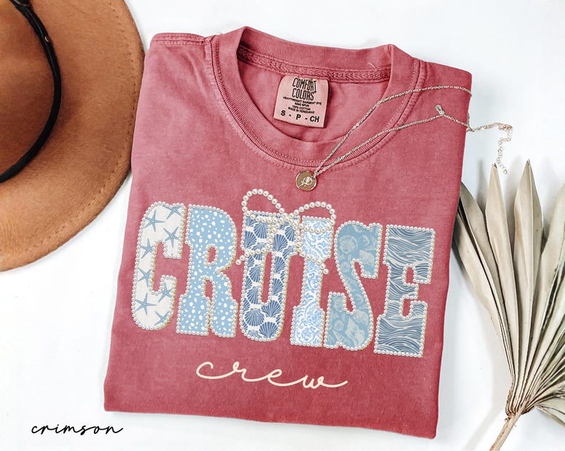 Puede incluir: Camiseta de color carmes&iacute; con las palabras "CRUISE crew". La palabra "CRUISE" es de color azul claro con un patr&oacute;n de conchas marinas y estrellas de mar. Un collar dorado con un colgante est&aacute; sobre la camiseta.