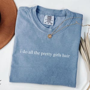 Może przedstawiać: Niebieska koszulka w kolorze dżinsu z białym napisem "i do all the pretty girls hair". Złoty naszyjnik z wisiorkiem i brązowy kapelusz r&oacute;wnież znajdują się na zdjęciu. Koszulka jest złożona i ma metkę.