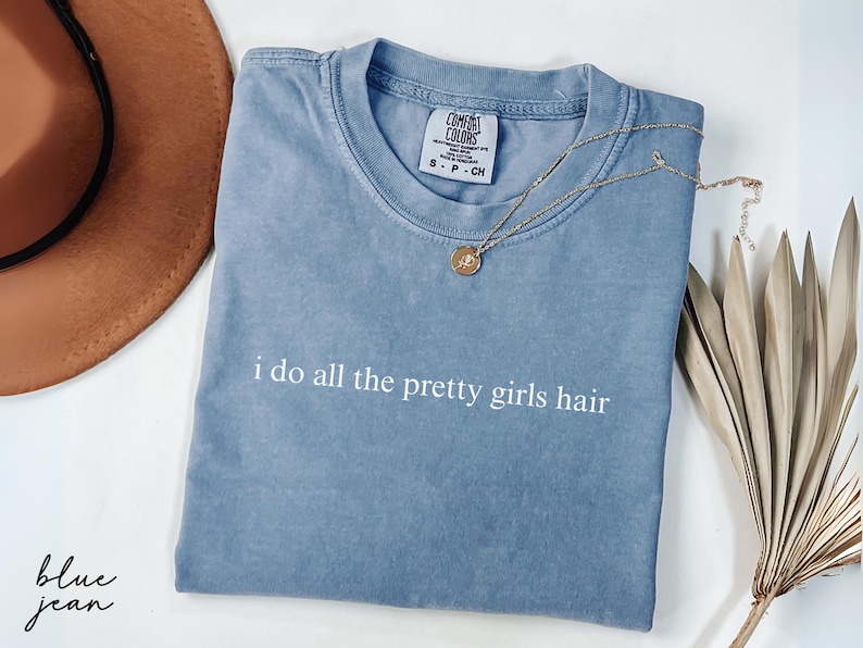 Może przedstawiać: Niebieska koszulka w kolorze dżinsu z białym napisem "i do all the pretty girls hair". Złoty naszyjnik z wisiorkiem i brązowy kapelusz r&oacute;wnież znajdują się na zdjęciu. Koszulka jest złożona i ma metkę.
