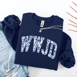 May include: Navy blue sweatshirt with the letters "WWJD" in white, filled with a blue floral pattern. A silver necklace with a small pendant rests on the sweatshirt. The sweatshirt is paired with a pair of blue jeans.