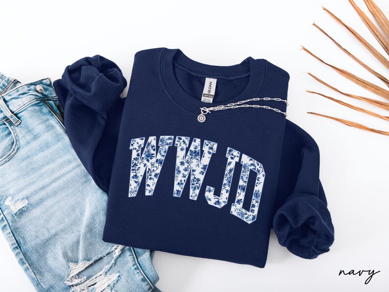 May include: Navy blue sweatshirt with the letters "WWJD" in white, filled with a blue floral pattern. A silver necklace with a small pendant rests on the sweatshirt. The sweatshirt is paired with a pair of blue jeans.