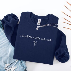 May include: A navy blue sweatshirt with the embroidered text "i do all the pretty girls nails" and a bow. A silver necklace with a circular pendant rests on the sweatshirt. Light blue denim jeans are visible on the left.