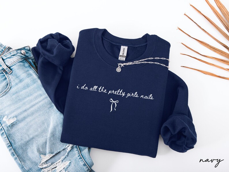 May include: A navy blue sweatshirt with the embroidered text "i do all the pretty girls nails" and a bow. A silver necklace with a circular pendant rests on the sweatshirt. Light blue denim jeans are visible on the left.