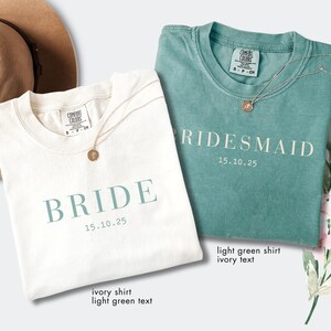 May include: Two t-shirts, one ivory and one light green, are displayed. The ivory shirt has "BRIDE" and "15.10.25" in light green text. The light green shirt has "RIDESMAID" and "15.10.25" in ivory text. Each shirt has a gold necklace.