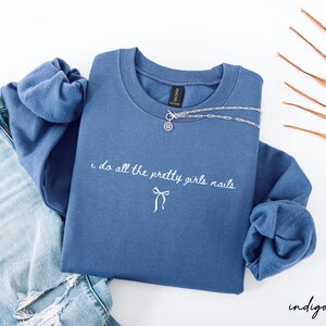 May include: Indigo blue sweatshirt with the text "i do all the pretty girls nails" in white script. A silver necklace with a pendant is draped across the top. The sweatshirt is paired with light blue denim jeans.