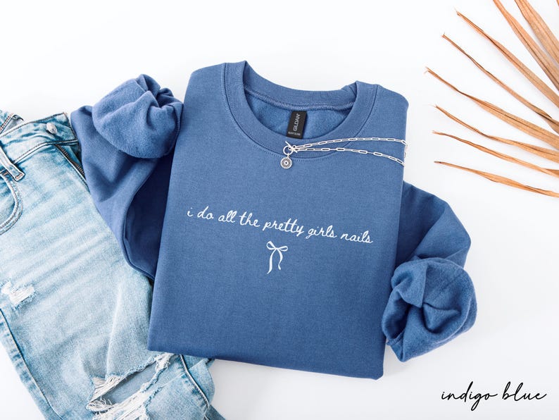 May include: Indigo blue sweatshirt with the text "i do all the pretty girls nails" in white script. A silver necklace with a pendant is draped across the top. The sweatshirt is paired with light blue denim jeans.