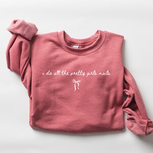 May include: A dusty rose sweatshirt with the text "i do all the pretty girls nails" in white script. A white bow graphic is below the text. The sleeves are rolled up, and the sweatshirt is on a white background.