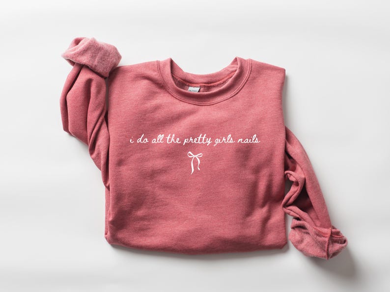 May include: A dusty rose sweatshirt with the text "i do all the pretty girls nails" in white script. A white bow graphic is below the text. The sleeves are rolled up, and the sweatshirt is on a white background.
