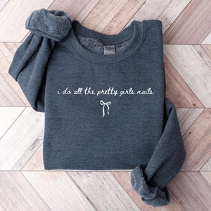 May include: A dark gray sweatshirt with the phrase "i do all the pretty girls nails" in white script, along with a small bow graphic. The sweatshirt is folded on a light brown and white wooden surface.